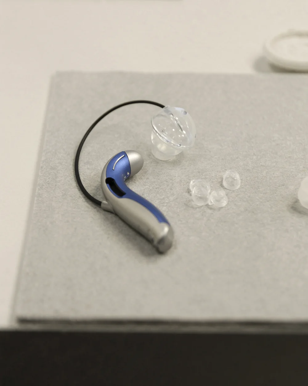 A single receiver in canal hearing aid lying on a grey felt pad beside three silicone domes and a wax filter tool, shown close so its size reads.