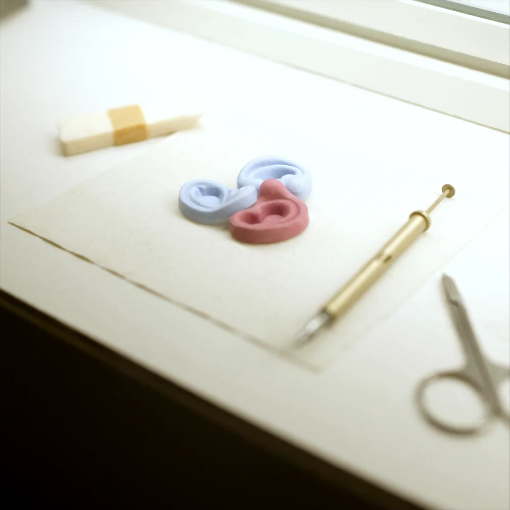 Three pairs of silicone ear impression castings in pale blue and pink resting on a white bench mat beside a syringe gun and curved scissors.