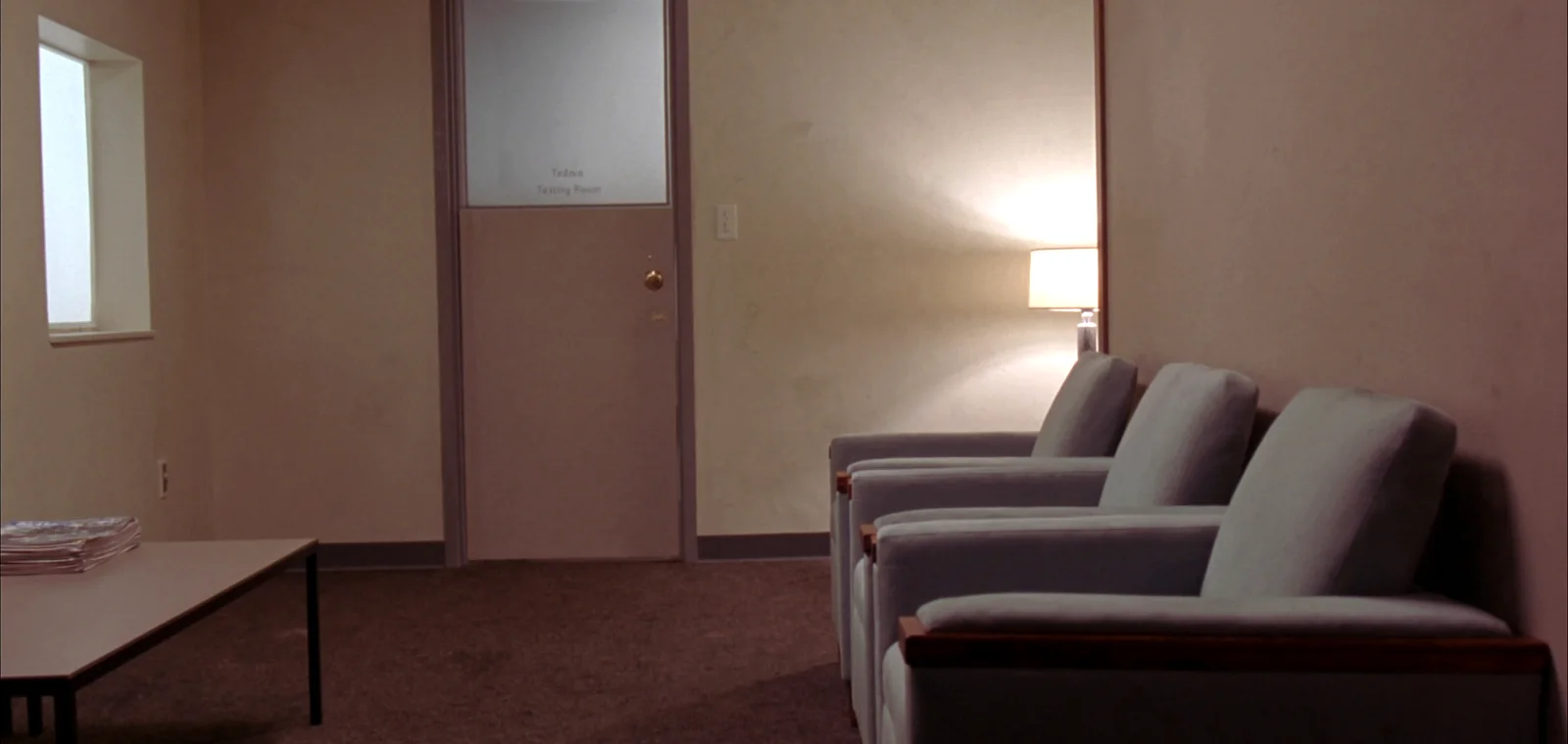 A small carpeted waiting room with four armchairs against a beige wall, a lamp, and a closed frosted glass door through to the testing room.